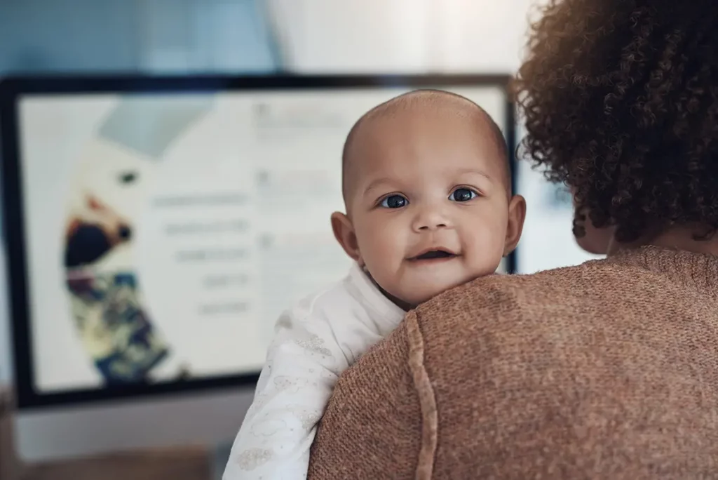 Un bébé souriant dans les bras de sa maman, avec un écran d’ordinateur flou en arrière-plan