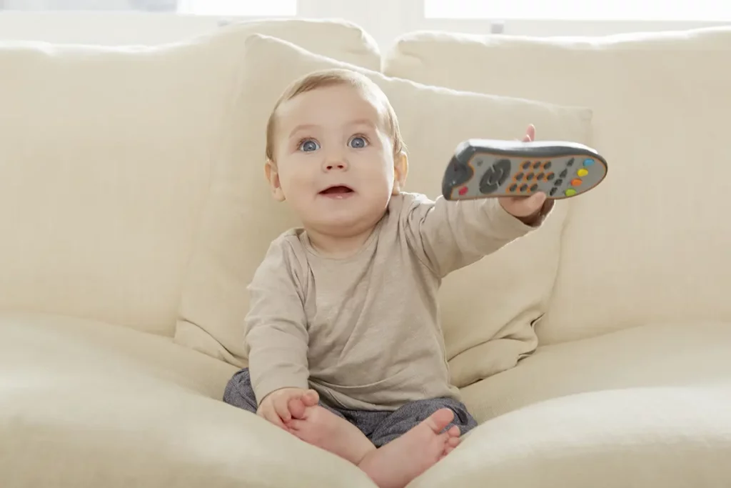 Un bébé assis sur un canapé beige tient une télécommande à une main en la pointant vers l’avant, les yeux écarquillés avec curiosité