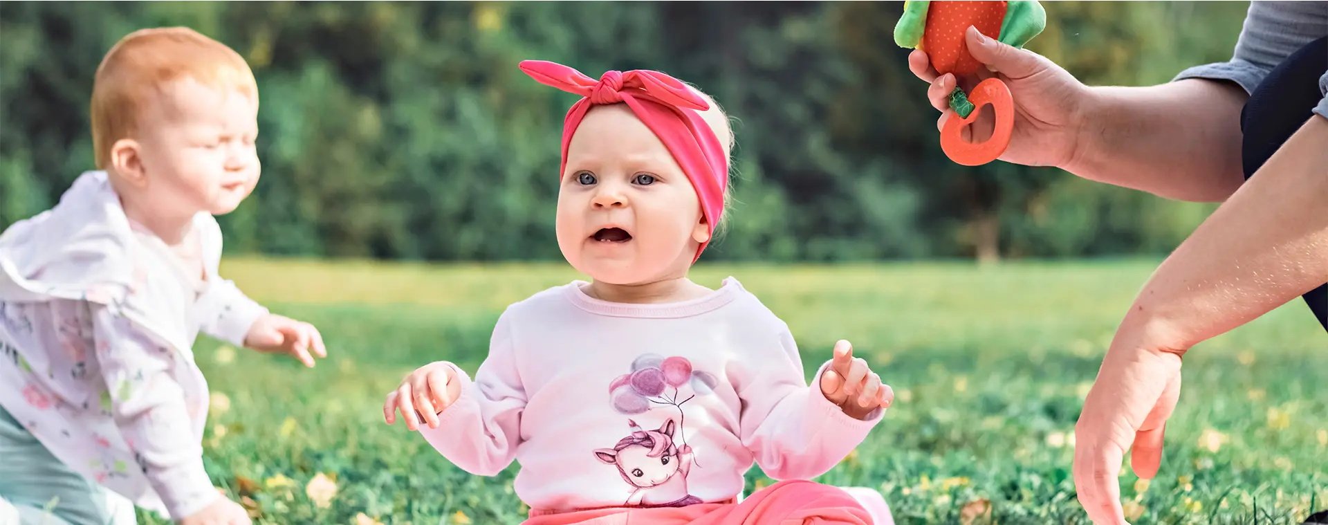 Deux bébés jouent dans un parc, accompagnés d’une nounou qui leur tend un jouet