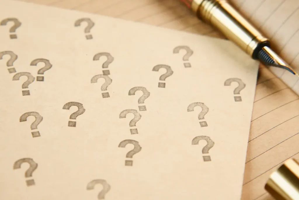 Feuille beige recouverte de points d’interrogation imprimés, avec un stylo plume doré posé à côté sur un carnet ligné, symbolisant les questions autour du projet de naissance