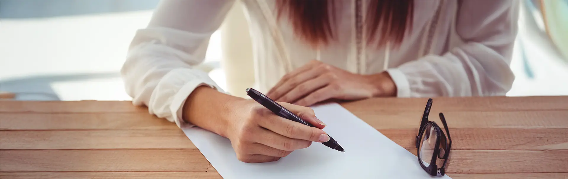 Gros plan sur une femme en haut blanc assise à une table en bois, tenant un stylo au-dessus d'une feuille blanche, ses lunettes posées à côté