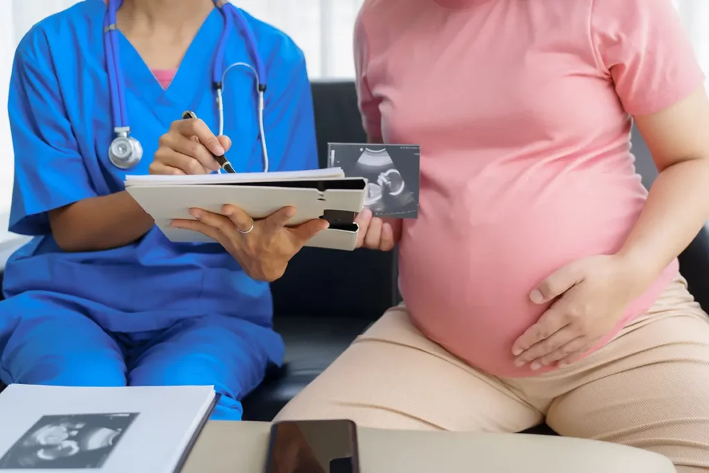 Femme enceinte en haut rose montrant son échographie à une sage-femme en blouse bleue qui prend des notes sur un dossier médical