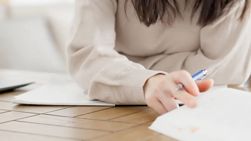 Gros plan sur une femme en pull beige, écrivant avec un stylo sur des feuilles posées sur une table en bois clair