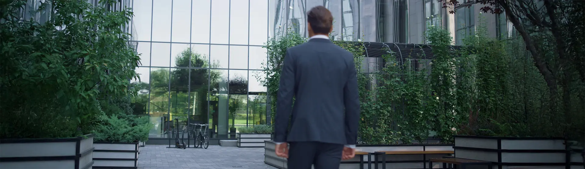 Un homme en costume vu de dos marche vers l’entrée d’un immeuble de bureaux modernes entouré de verdure, symbolisant la reprise du travail ou le retour au bureau