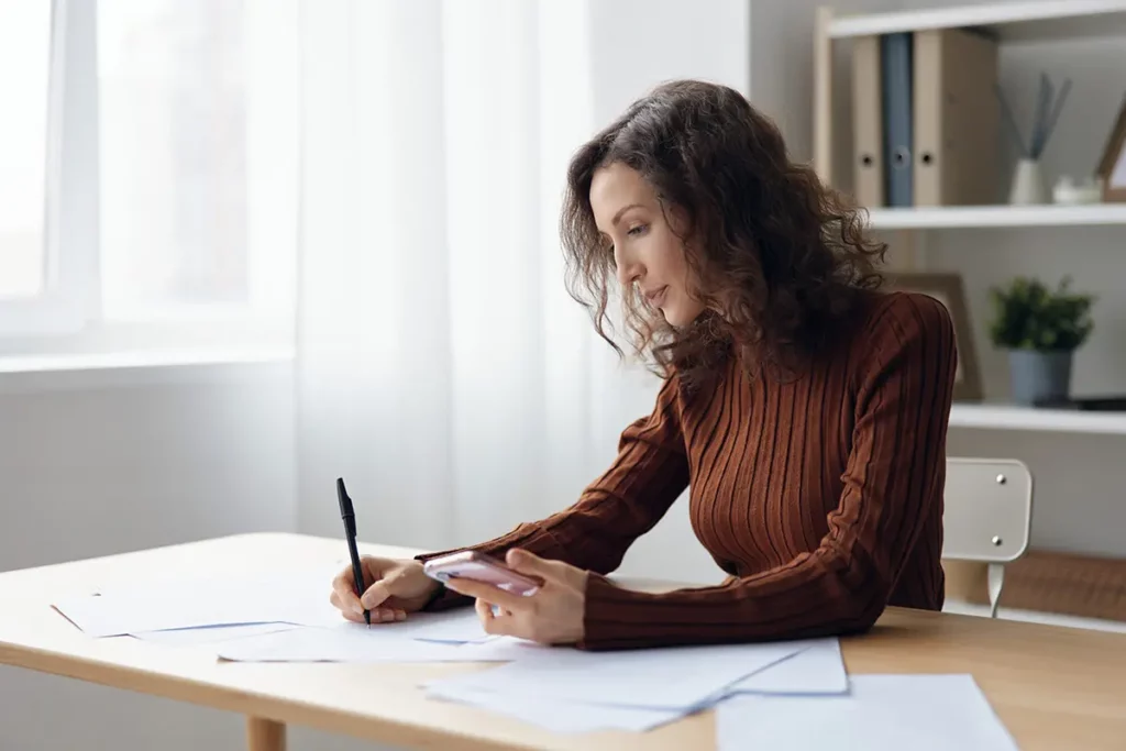 Femme concentrée remplissant des documents administratifs à une table, stylo à la main et téléphone dans l’autre, entourée de papiers
