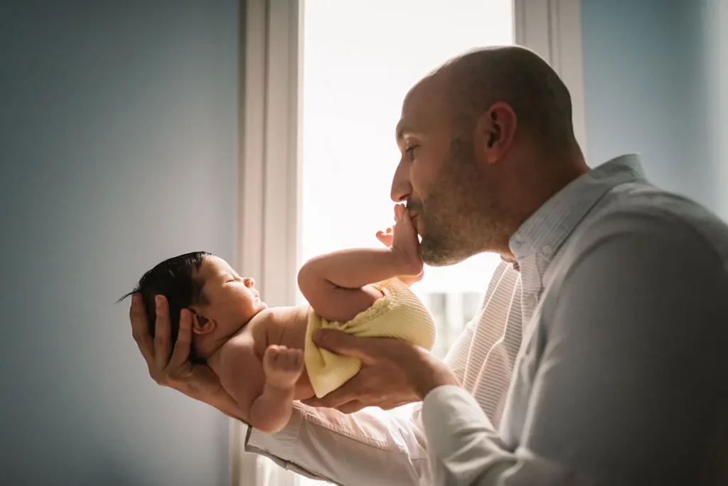 Un père souriant tient tendrement son bébé dans les bras et embrasse son pied, devant une fenêtre baignée de lumière naturelle