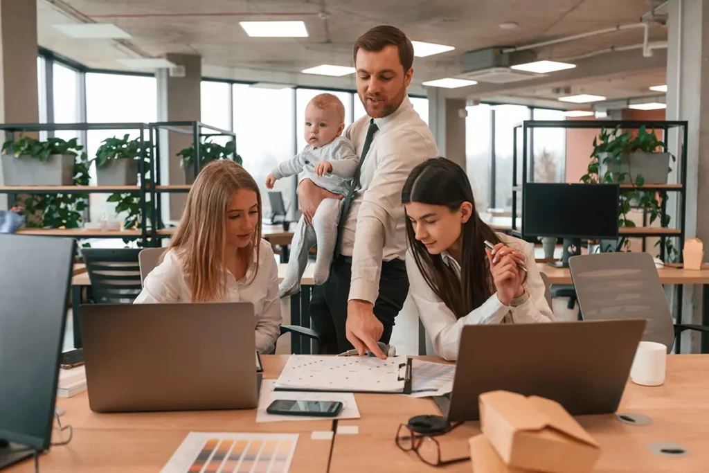Un père en tenue professionnelle tient son bébé dans les bras tout en discutant avec deux collègues autour d’une table de travail dans un bureau moderne, illustrant la conciliation entre vie professionnelle et vie familiale
