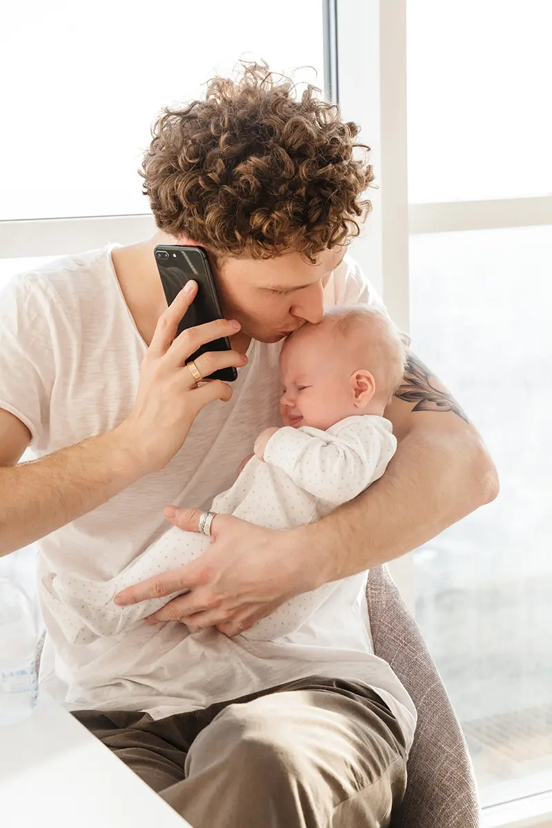 Jeune papa tenant tendrement son bébé dans ses bras tout en passant un appel téléphonique à sa mutuelle pour organiser la couverture santé de son enfant
