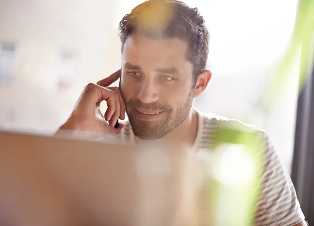 Homme souriant au téléphone tout en consultant son ordinateur portable, organisant les démarches de mutuelle pour son futur bébé