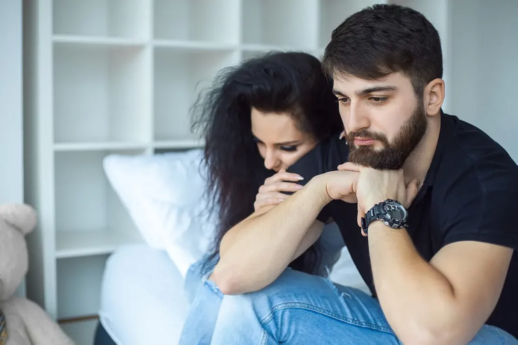 Un couple assis sur un lit, le regard préoccupé et triste, symbolisant les tensions ou la détresse émotionnelle que peuvent traverser certains parents