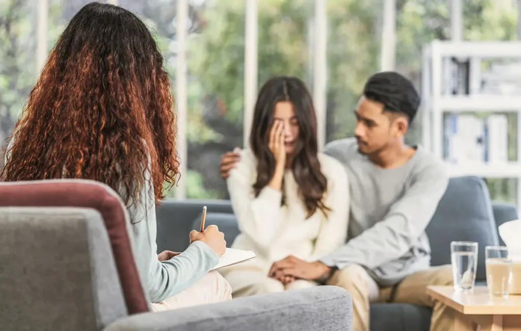 Un couple en détresse émotionnelle assis sur un canapé, face à une thérapeute prenant des notes, illustrant une séance de thérapie de couple dans un cadre apaisant
