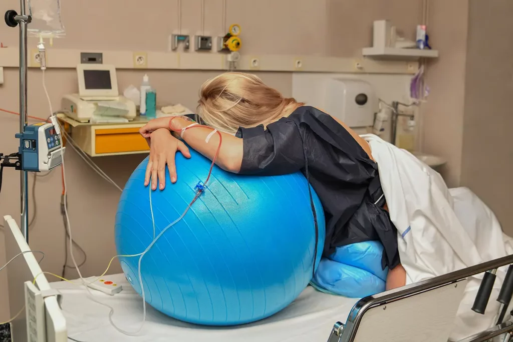 Femme enceinte en travail à la maternité, s’appuyant sur un gros ballon d’exercice bleu pour soulager ses contractions en variant les positions