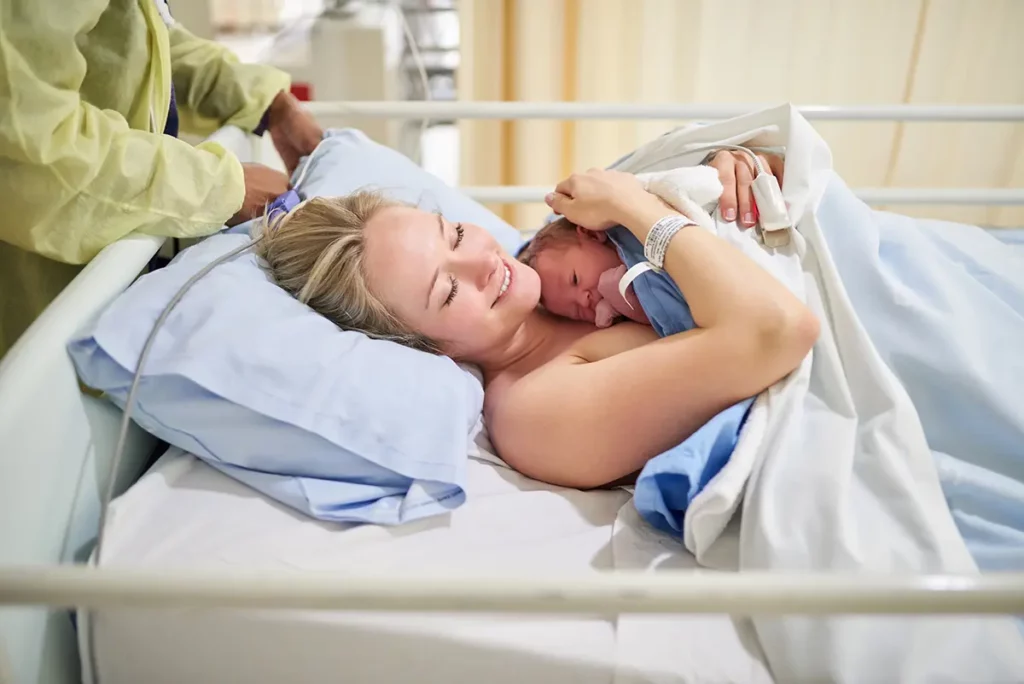 Maman souriante allongée dans son lit d’hôpital avec son nouveau-né blotti contre elle après l’accouchement