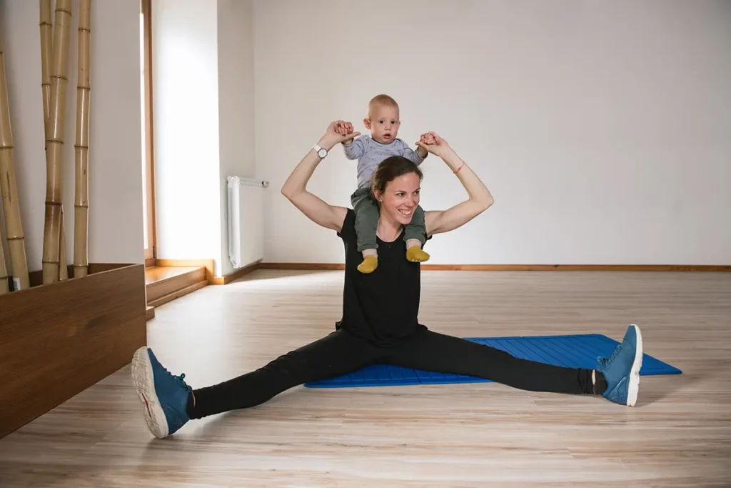 Maman en tenue de sport assise en grand écart sur un tapis, souriante, portant son bébé sur ses épaules tout en tenant ses mains, illustrant un moment de gym douce et complice