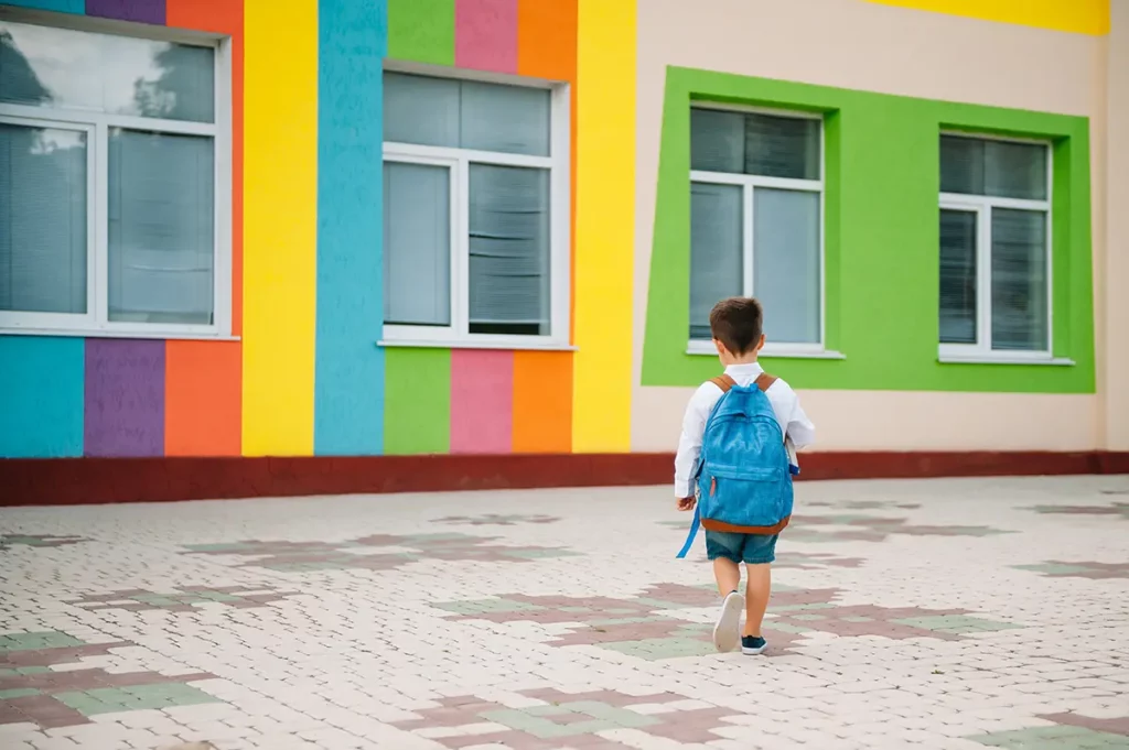 Petit garçon de 3 ans avec un sac à dos bleu marchant vers l’école maternelle colorée