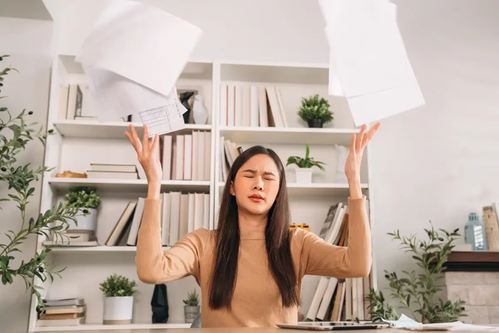 Jeune maman débordée assise à son bureau, les yeux fermés, lançant des papiers en l'air par frustration ou lassitude, illustrant le stress du quotidien