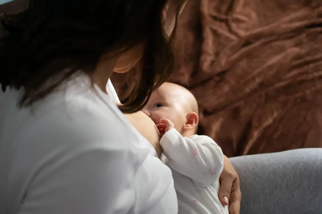 Jeune maman en chemise blanche allaitant tendrement son bébé blotti contre elle, illustrant un moment d’allaitement calme et intime sur un canapé