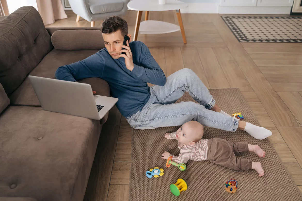 Un jeune père assis au sol, téléphone à l’oreille et ordinateur portable sur le canapé, tout en s’occupant de son bébé allongé près de lui sur un tapis de jeu avec des jouets colorés