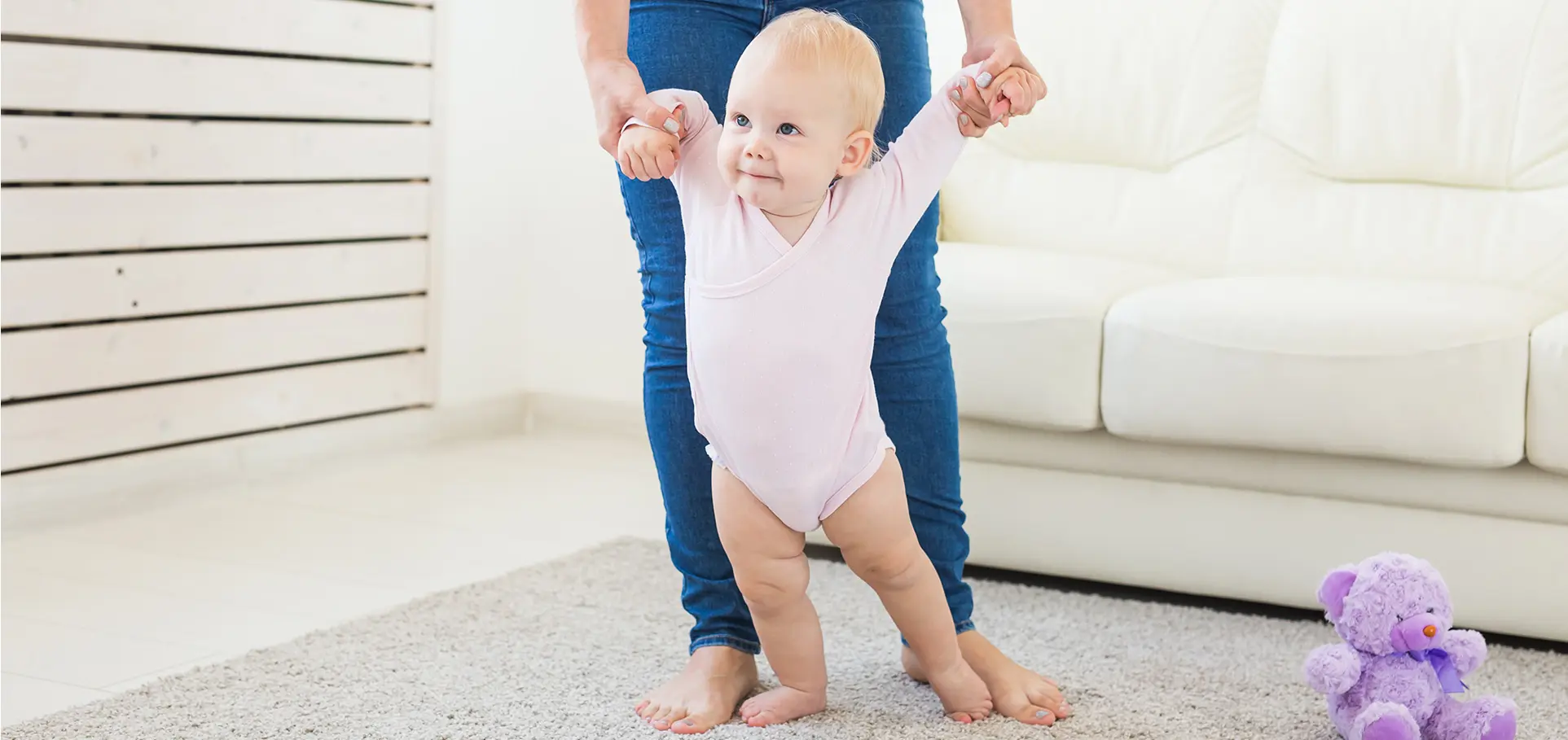 Maman qui tient sa fille par les bras pour lui apprendre à marcher sur un tapis.