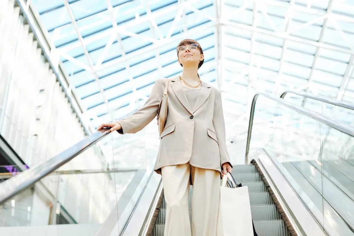 Une femme élégamment vêtue descend un escalator dans un bâtiment moderne et lumineux, tenant un sac à la main, symbole de son retour à la vie professionnelle après la maternité