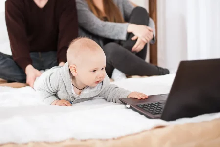Bébé attrape le PC Un bébé allongé sur un lit tend la main vers un ordinateur portable, sous le regard attendri de ses parents assis en arrière-plan