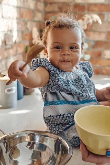 Bébé sur le plan de travail avec un ustensile en main Un bébé assis sur un plan de travail de cuisine, tenant une cuillère en bois et un bol, avec un grand sourire espiègle