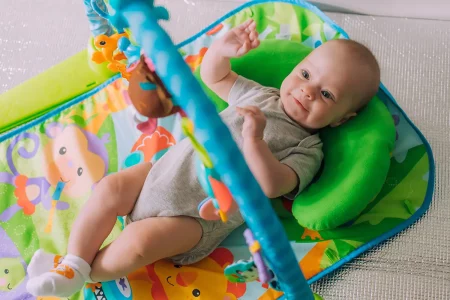 Bébé sur tapis de développement Un bébé allongé sur un tapis d’éveil coloré avec des jouets suspendus, souriant et tendant les bras pour jouer, appuyé sur un coussin vert