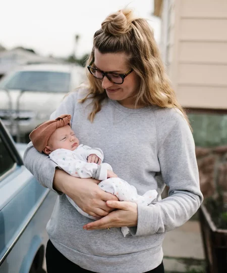 Femme avec bébé retour maison Jeune maman en sweat gris tenant tendrement son nourrisson emmailloté dans ses bras devant la maison, souriante et attentionnée lors du retour à la maison après la maternité