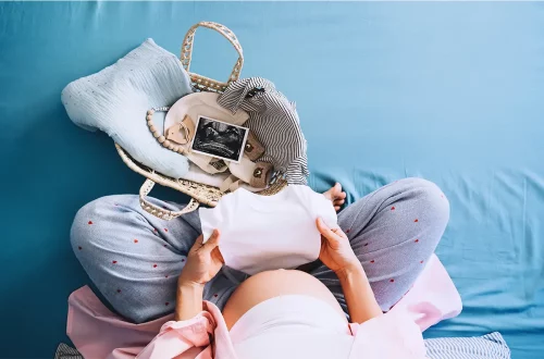 Femme enceinte qui prépare l’arrivée de bébé Vue de dessus d'une femme enceinte assise en tailleur sur un lit bleu, tenant un body blanc pour bébé et préparant un sac de naissance rempli de vêtements et d’une échographie