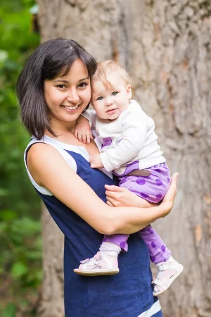 Une jeune femme souriante tenant son bébé dans les bras, tous deux regardant l’objectif, en extérieur devant un arbre