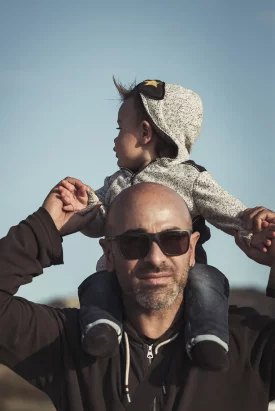 Un homme portant des lunettes de soleil tient son bébé assis sur ses épaules, profitant d'une journée en plein air sous un ciel bleu