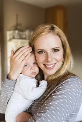 Une jeune femme souriante tient tendrement son bébé dans ses bras, tous deux habillés de vêtements confortables, dans un intérieur chaleureux