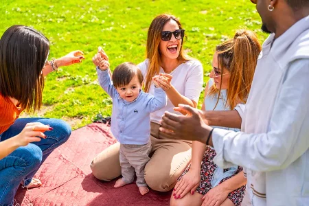 Groupe de parents assis sur une couverture au parc, souriants et jouant avec un bébé qui apprend à marcher