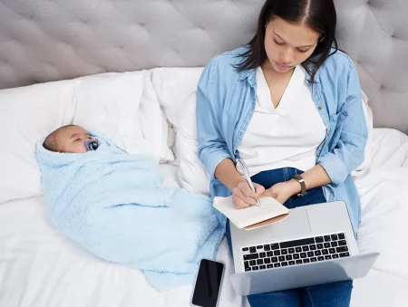 Maman travaillant sur son ordinateur portable depuis le lit avec son bébé emmailloté à ses côtés