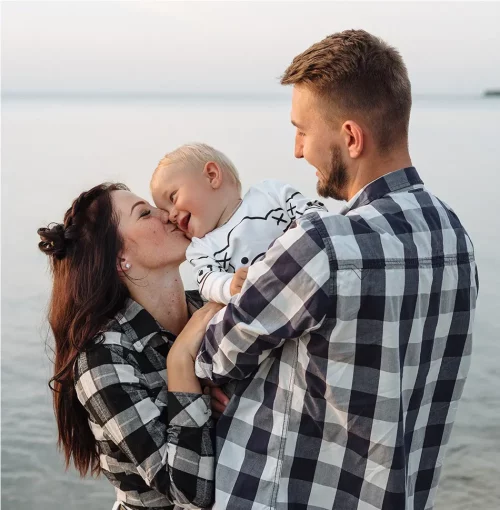 Un couple souriant tient et embrasse leur bébé blond au bord de la mer, tous habillés de façon coordonnée en chemises à carreaux, créant une scène de tendresse familiale au coucher du soleil